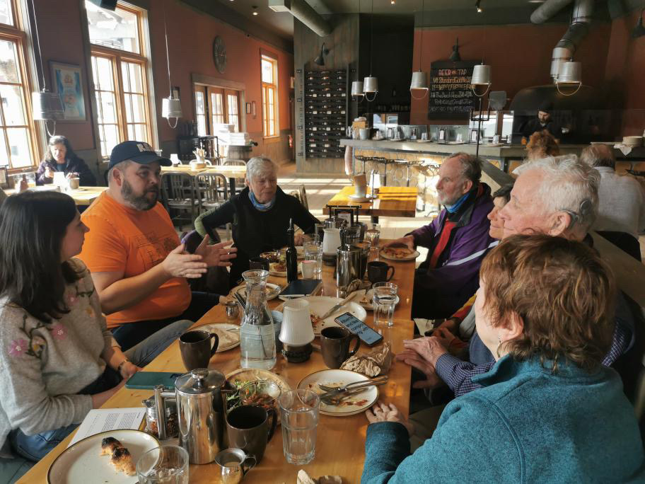 A dozen people seated around a long table in a Nelson cafe, mid-conversation over coffee and breakfast