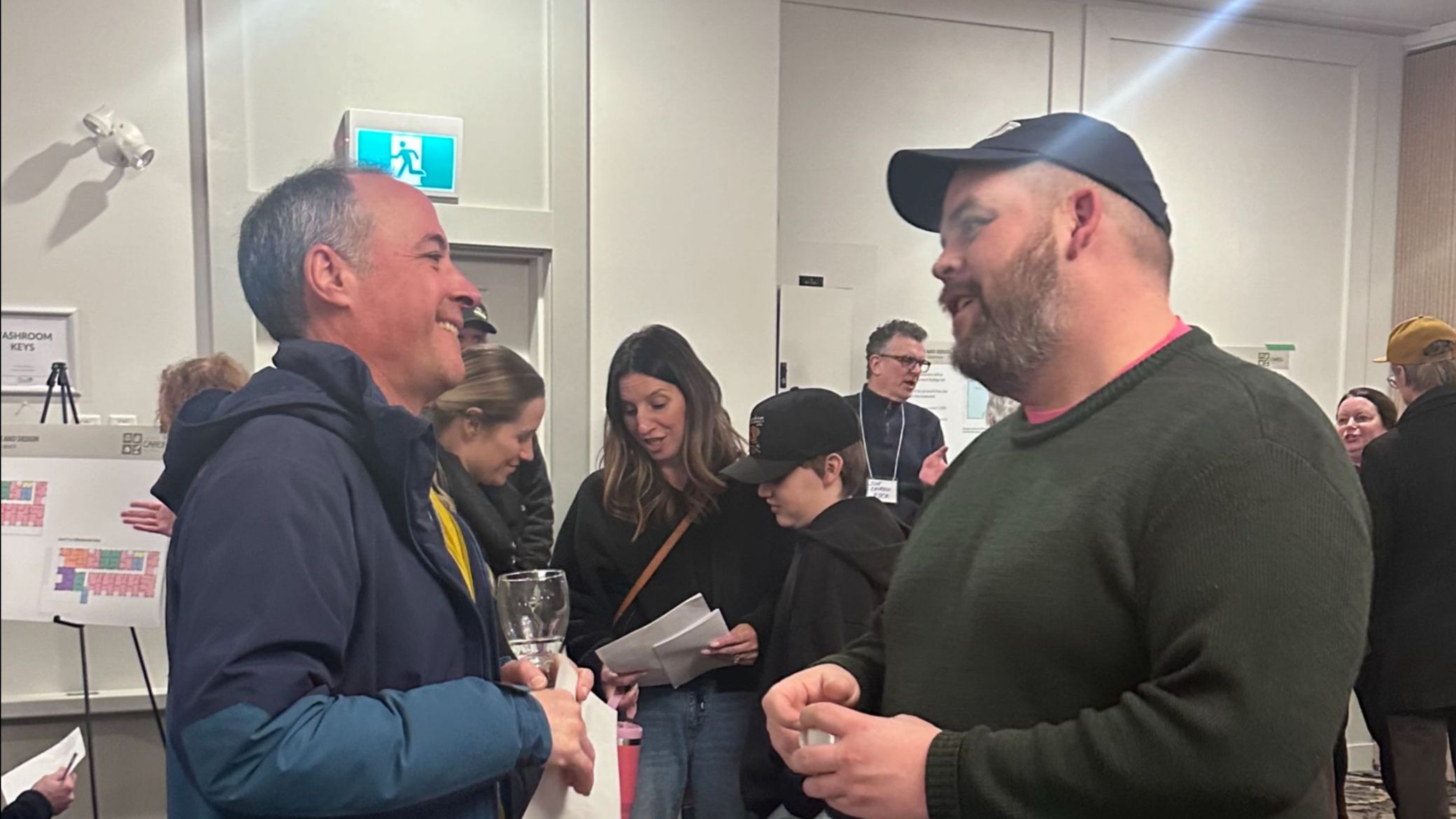 Two people talking face to face at an indoor community event, with a crowd gathered behind them