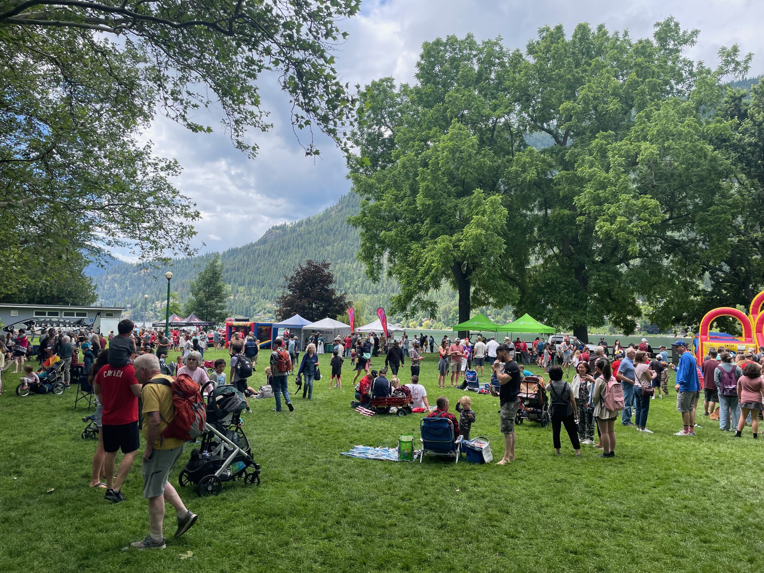 A crowd spread across a lakeside park in Nelson on a summer day, with forested mountains behind